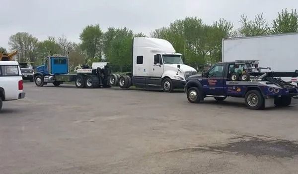 A white semi-truck being towed by a blue tow truck on a grey parking lot.