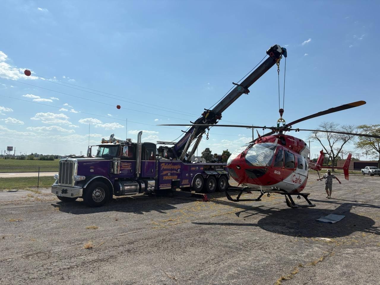 Purple tow truck lifting a red and white helicopter outdoors under a blue sky.