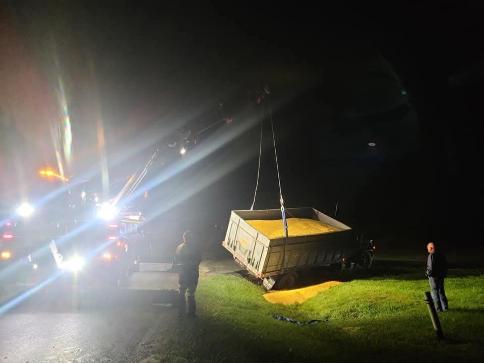 A tow truck lifts a tipped trailer filled with yellow grain at night. A few people watch.