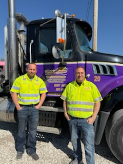 Two men in neon shirts stand in front of a tow truck.