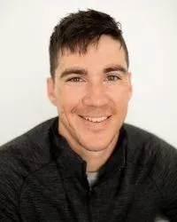 Smiling person with short dark hair in a dark zip-up jacket against a light background