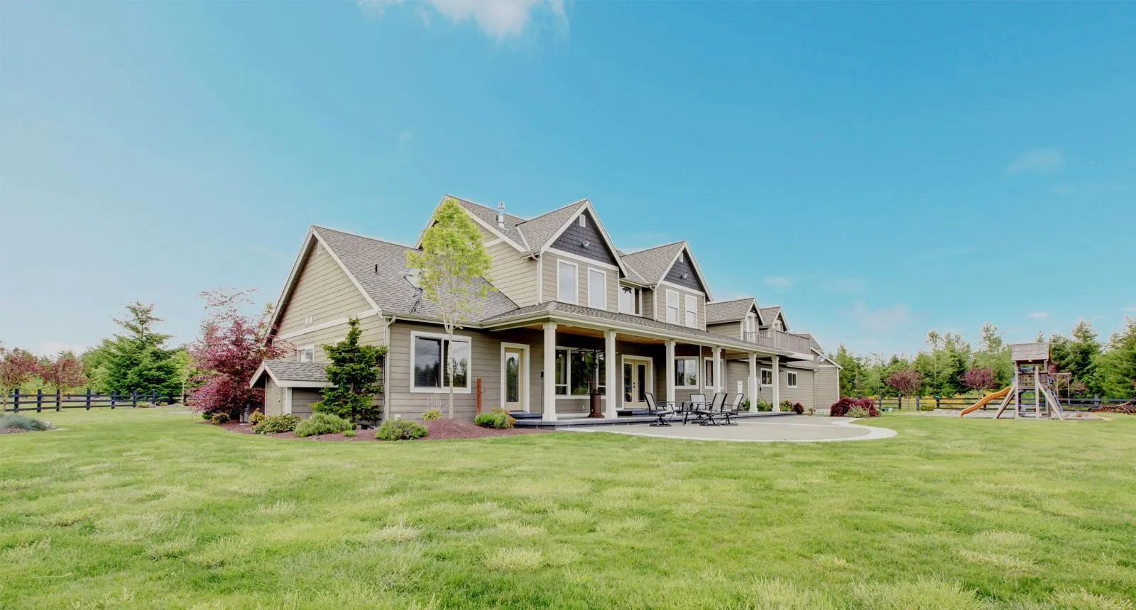 Large gray house with a covered porch and wide green lawn under a bright blue sky
