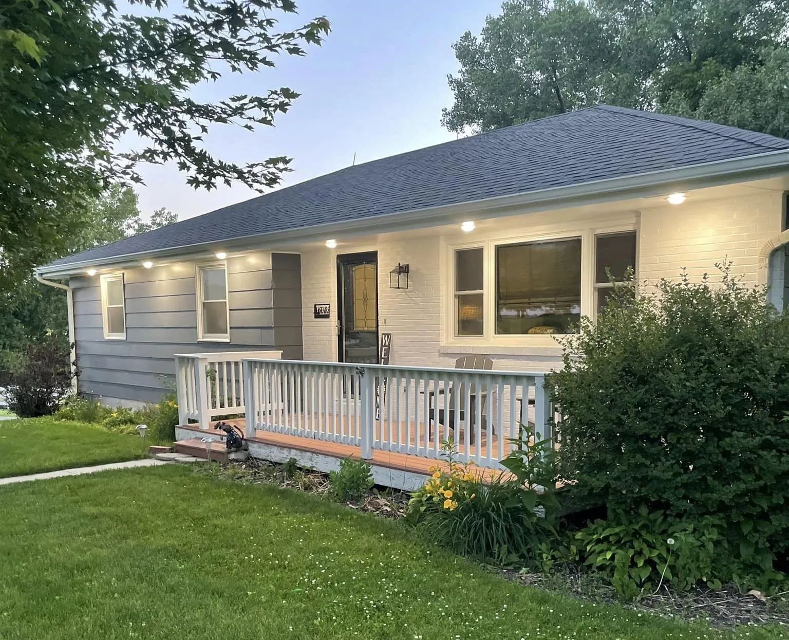 Single-story gray house with a front porch, lit exterior, and green lawn at dusk