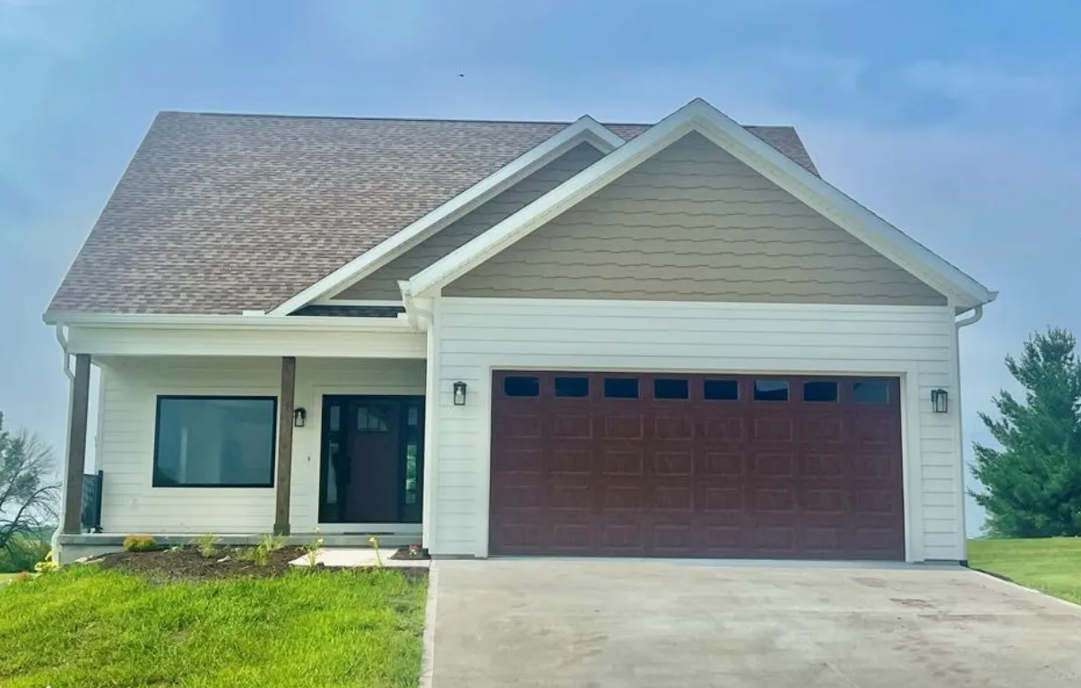 Single-story suburban house with beige siding, brown garage door, and a concrete driveway under a partly cloudy sky