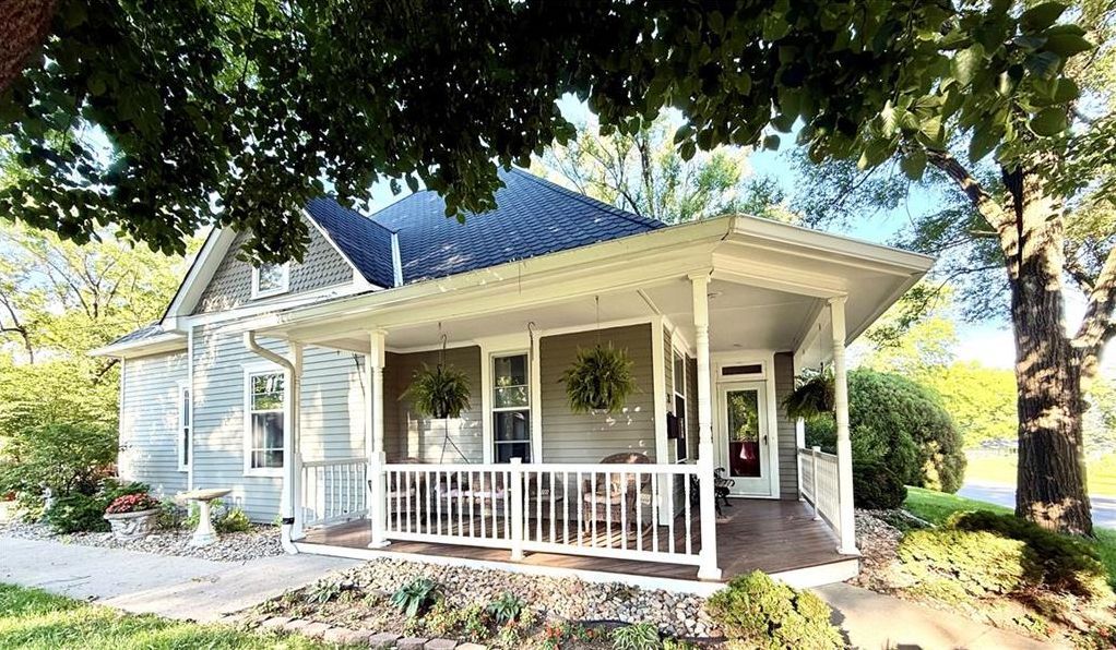 White farmhouse with a wraparound porch and blue roof surrounded by trees and garden beds