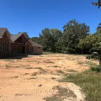 A large house is sitting in the middle of a dirt field surrounded by trees.