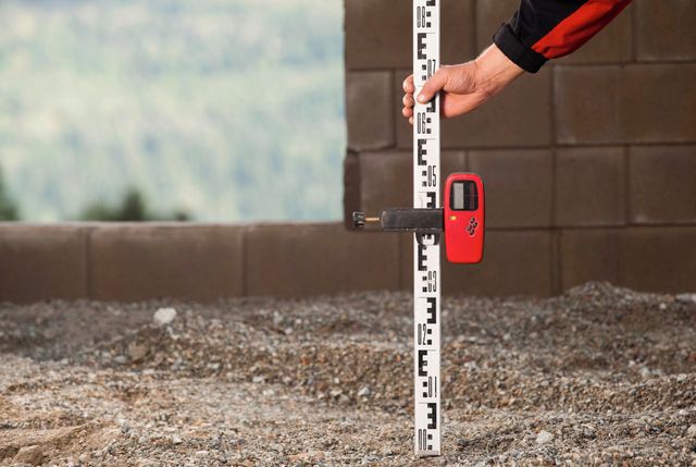 Person using a leveling rod with a red digital reader on a construction site.