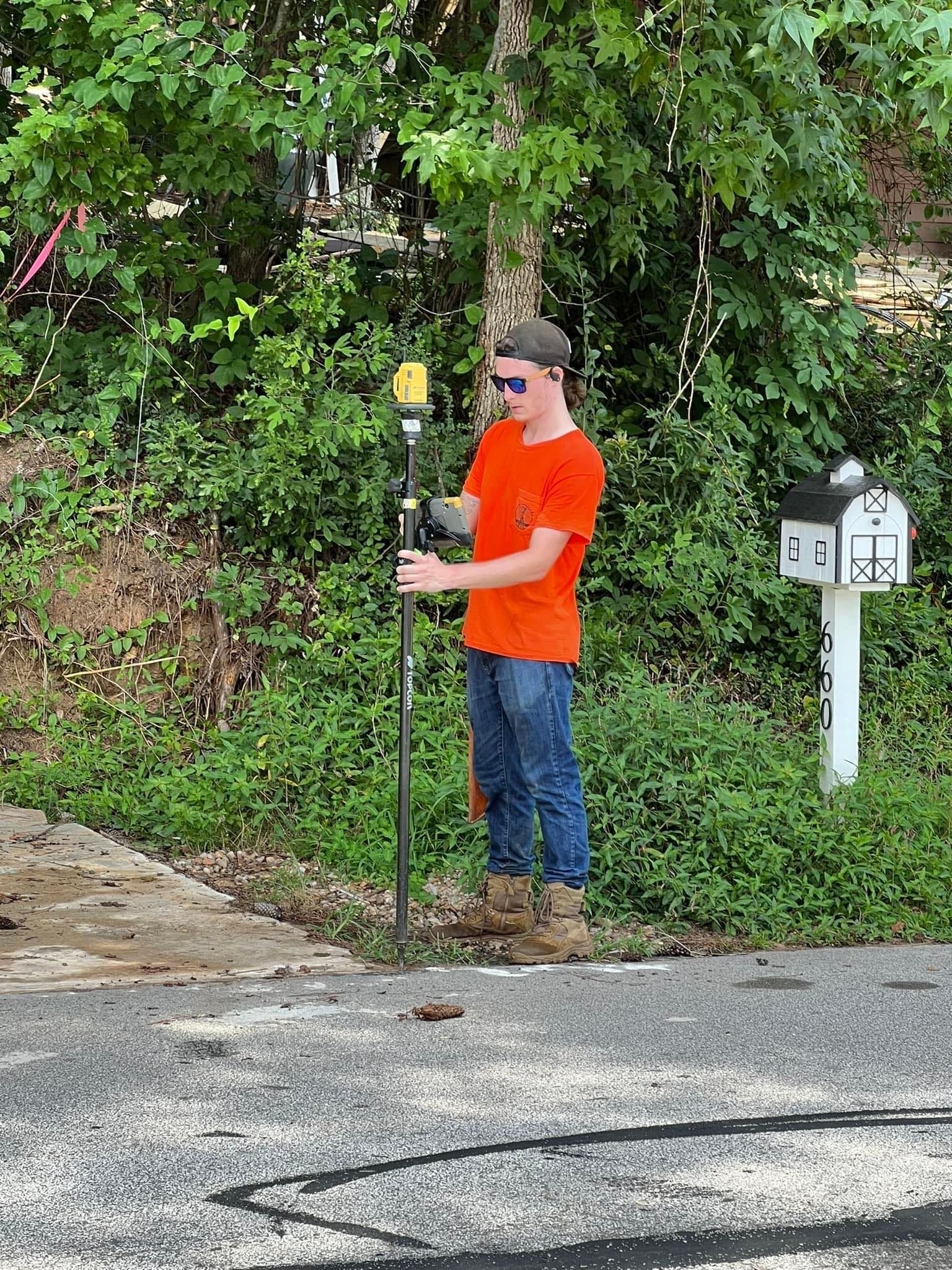 Person in orange shirt holds a surveying pole.  He stands near a tree, mailbox, and dense foliage.