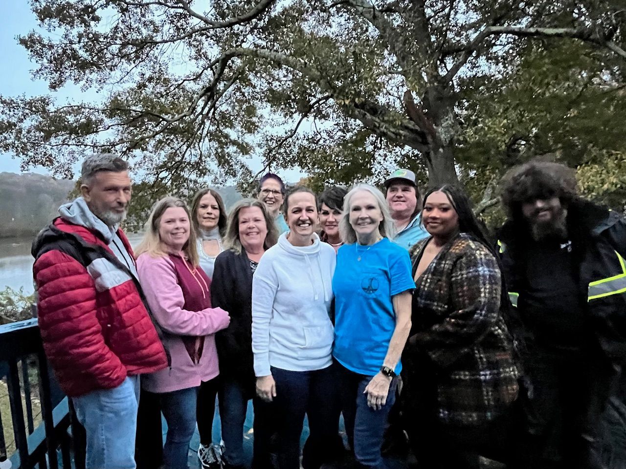 Group of people, mostly women, smile for a photo outdoors near water. Overcast day.
