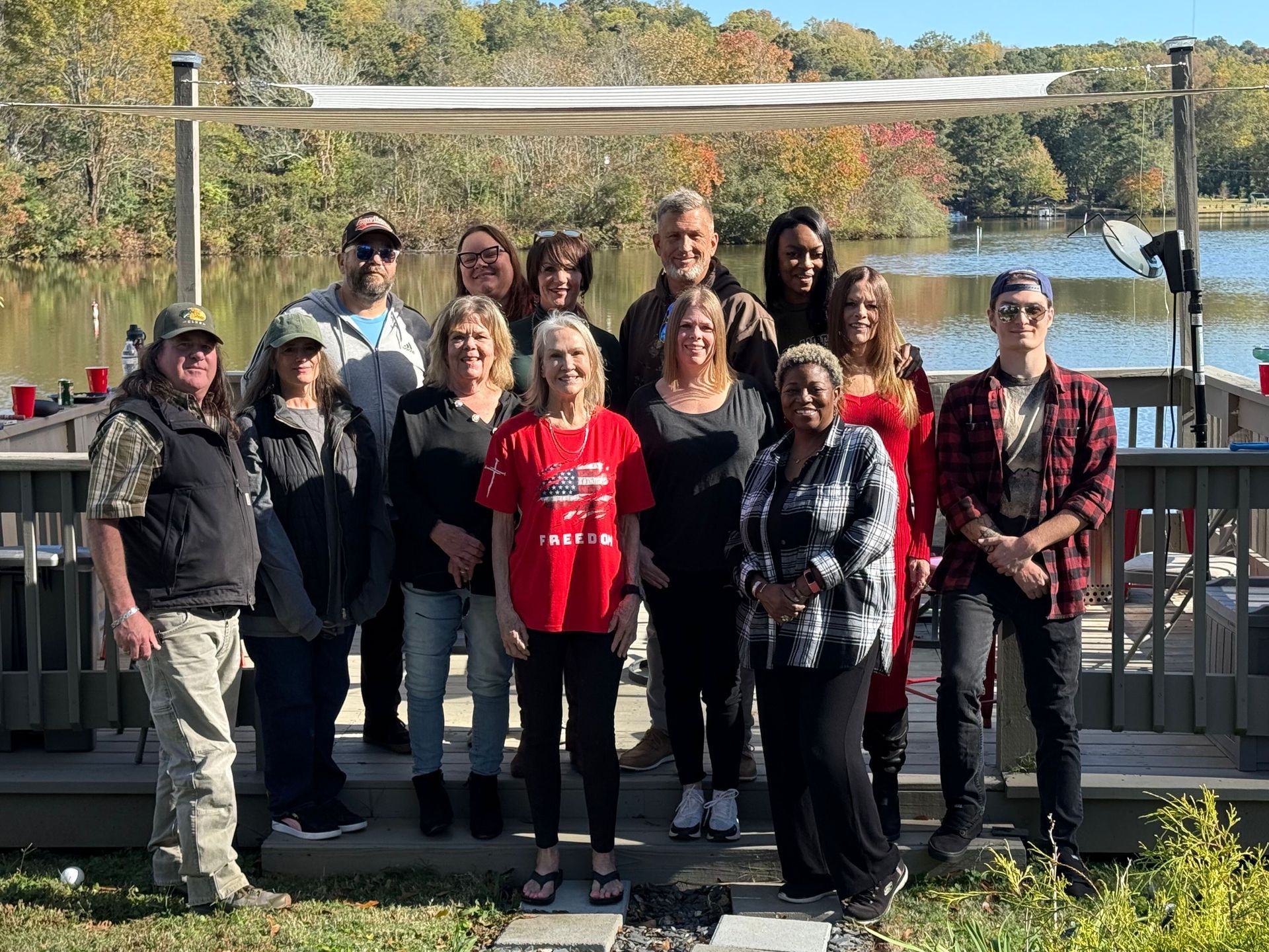 Group of people posing outdoors by a lake, autumn foliage, under a gazebo.
