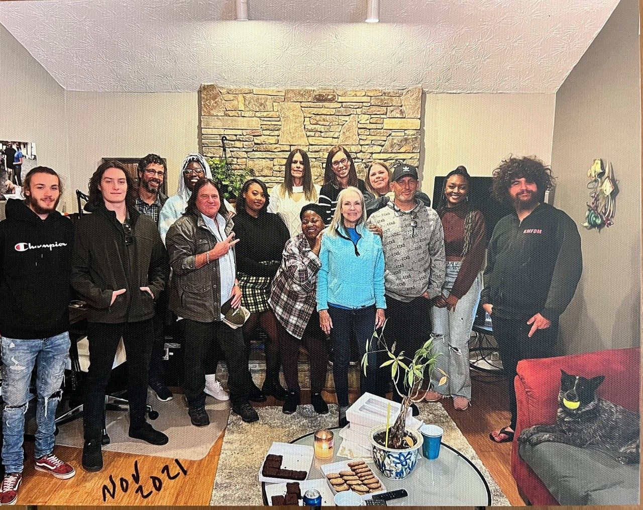 Group of people pose inside a room with a stone fireplace. Some stand, others sit. A coffee table has items on it.
