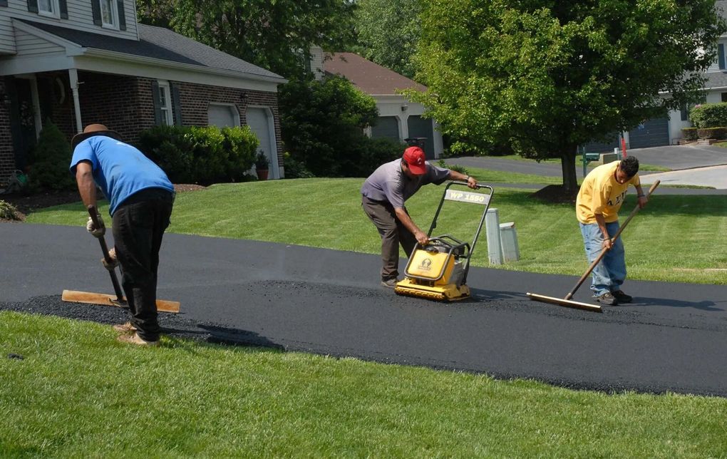 Three workers paving a driveway: one smoothing with a tool, one using a plate compactor, and one spreading asphalt.