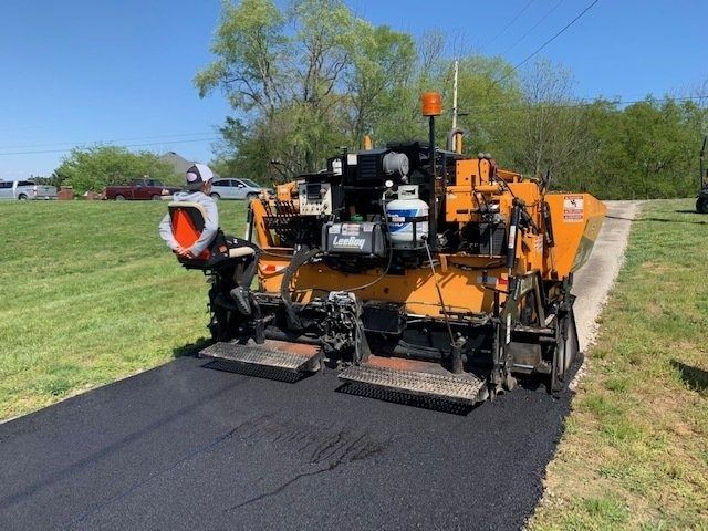 An orange asphalt paver lays a smooth strip of fresh black pavement on a sunny day in a grassy, outdoor setting.
