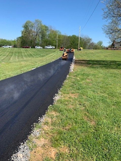 A steamroller flattens freshly laid black asphalt on a driveway through a grassy field under a bright blue sky.