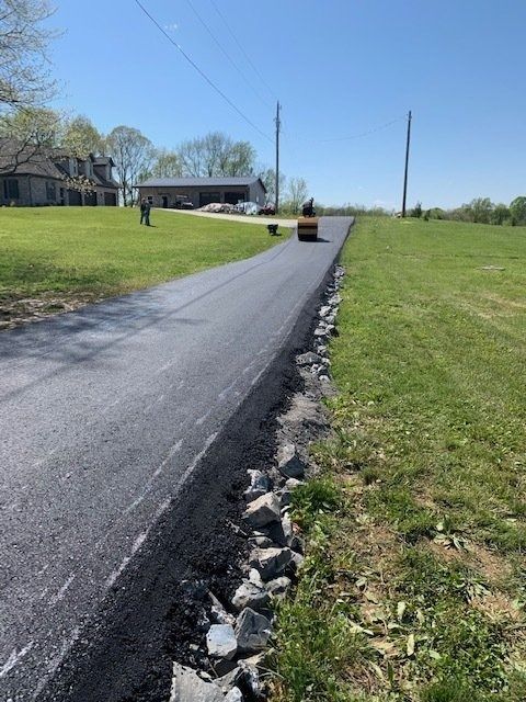 A construction vehicle rolls fresh asphalt on a driveway in a rural, grassy setting.