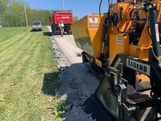A bright yellow road paver lays asphalt on a rural road with a red dump truck and worker visible in the distance.