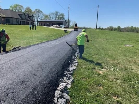 Workers in high-visibility vests pave a driveway with asphalt next to a grassy field on a sunny day.