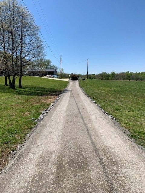 A long, paved driveway stretches through a green field toward a house under a clear, bright blue sky.