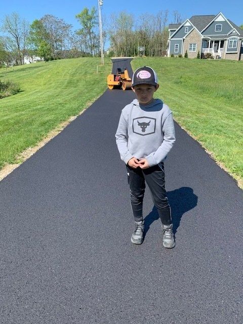 A young person wearing a grey hoodie and cap stands on a newly paved asphalt driveway in front of a house and machinery.