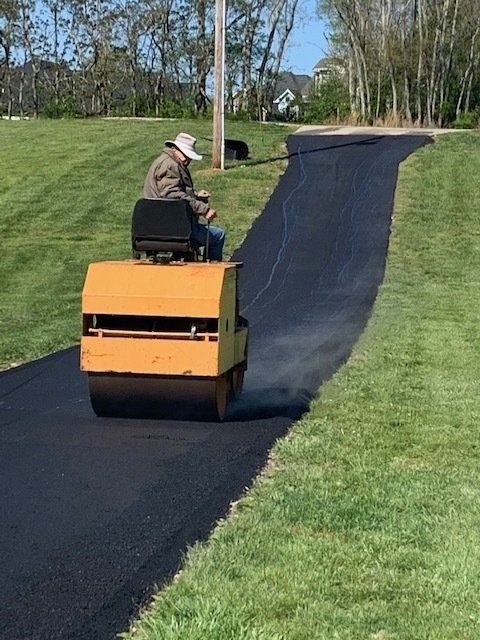 A person drives a yellow steamroller over a newly paved asphalt driveway on a grassy hill.