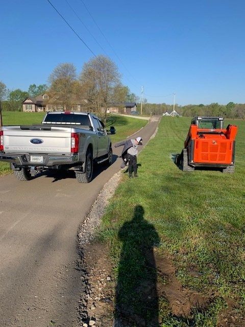 A person in work clothing stands on a grassy roadside between a silver pickup truck and an orange skid-steer loader.