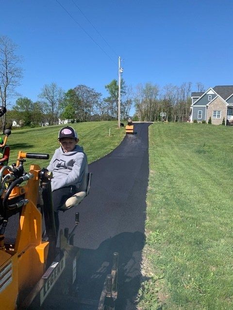 A person operating a yellow asphalt roller on a newly paved driveway leading toward a suburban house.