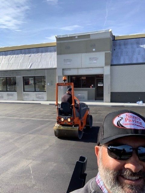A smiling man in a branded hat and sunglasses takes a selfie in front of a construction site with an orange road roller.