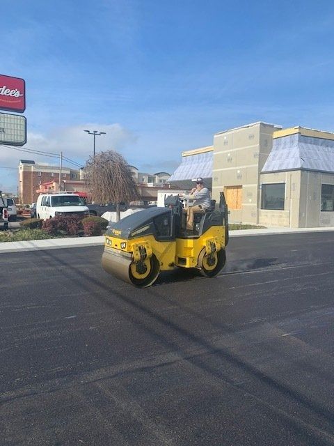 A yellow asphalt roller being driven on freshly paved black pavement in front of a commercial building.