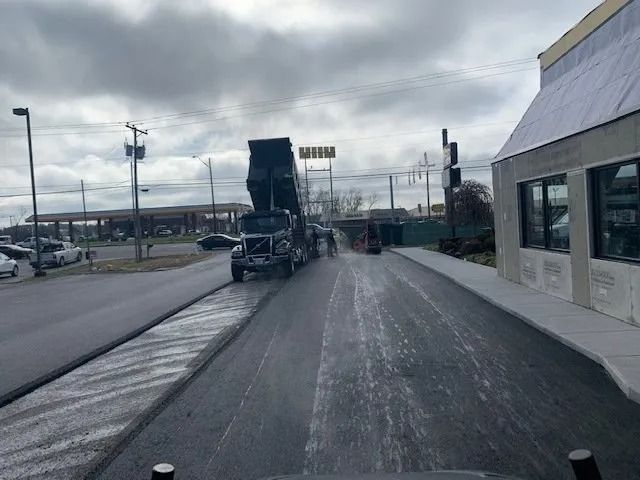 A dark-colored dump truck with its bed raised, driving on a newly paved road next to a building under cloudy skies.