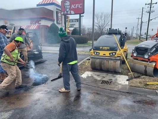 Workers in high-visibility vests and casual clothes repair asphalt in a parking lot near a KFC restaurant.