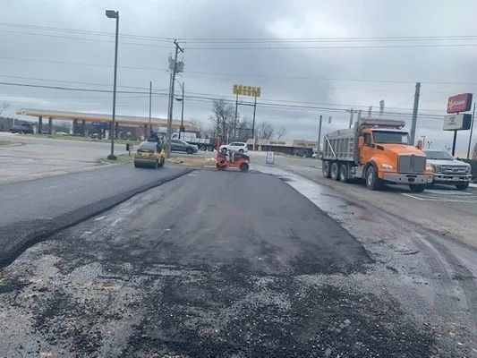 An orange construction roller levels freshly laid asphalt in a parking lot, with a dump truck and cars in the background.