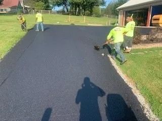 Four workers in yellow high-visibility shirts pave a long asphalt driveway on a sunny day near a building.