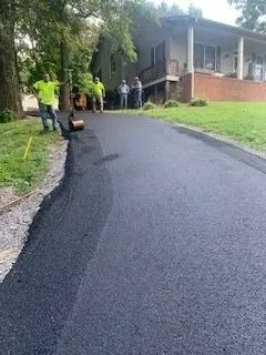 Workers pave a new asphalt driveway leading to a house, with a hand roller visible on the edge.