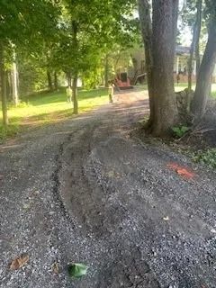 Two people stand on a gravel driveway near a house, with heavy equipment visible in the background under trees.