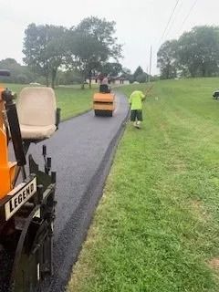 A worker in a neon yellow shirt stands beside a fresh asphalt path as a steamroller travels along it on a grassy hillside.