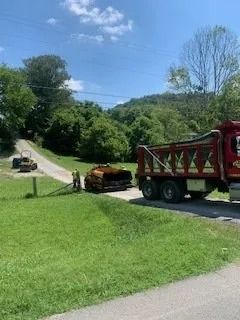 A red dump truck and a construction paver on a rural paved road during a bright, sunny day.