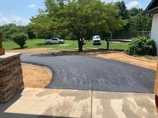 A newly paved asphalt driveway curves through a residential yard, leading toward a tree and parked white vehicles.