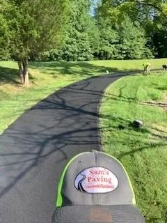 A person wearing a Sam's Paving hat stands on a freshly paved black asphalt path winding through a grassy park.