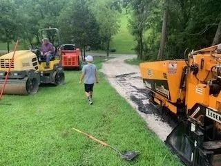 A child walks between a yellow road roller with an operator and a yellow asphalt paver on a rural gravel driveway.