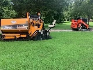 An orange LeeBoy 7000C asphalt paver sits on a grassy lawn next to a smaller orange tracked skid steer in the distance.