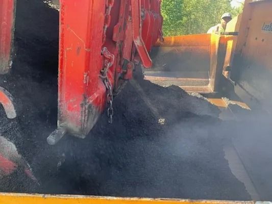 A red asphalt paver machine spreads hot, steaming black asphalt onto a road surface.