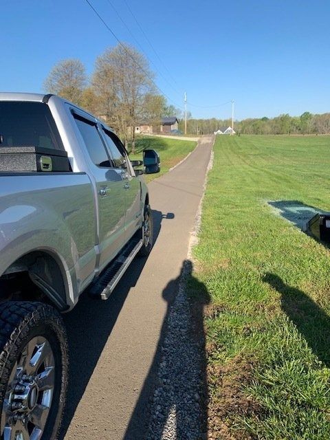 A silver truck parked on a narrow road next to a grass field on a sunny day, with a long shadow cast across the pavement.