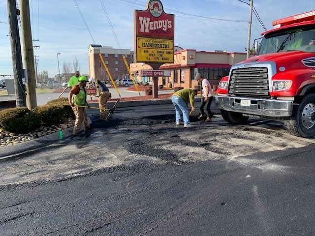 A crew of workers in neon safety vests repairs a patch of asphalt in a Wendy's parking lot near a red truck.