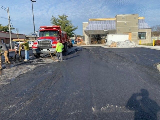Construction workers paving a parking lot in front of a building with a red dump truck nearby.