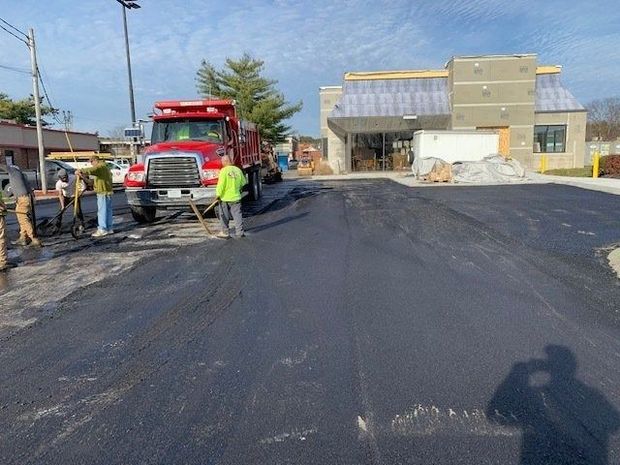 Workers paving a parking lot with a red dump truck in front of a commercial building under construction.