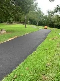 A newly paved black asphalt driveway winds through a green grassy lawn toward a distant orange construction roller.