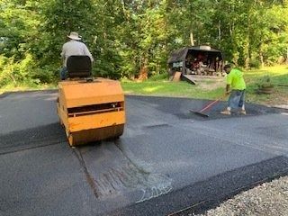 A worker operates a yellow steamroller to flatten fresh asphalt while another worker nearby spreads it with a rake.