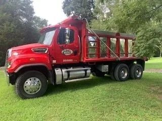 A bright red dump truck parked on a lush green lawn surrounded by trees.