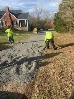 Two people in high-visibility yellow shirts rake fresh gravel onto a driveway in front of a brick house.