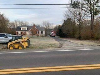 A yellow skid steer sits on a grassy lawn next to a driveway and a house, with a construction truck visible in the back.